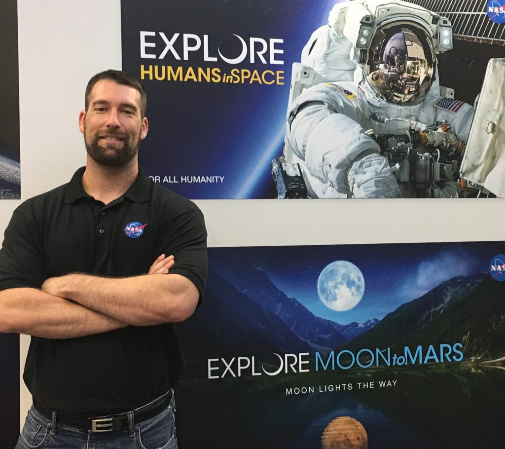 Matt Wittal smiles as he stands near an Artemis poster with "Explore Moon to Mars" and an astronaut's picture