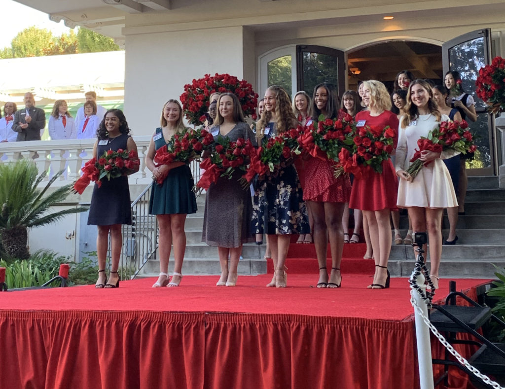The seven members of the 2020 Royal Court pose with red roses on red-carpeted platform behind Tournament House as White Suiters look on.