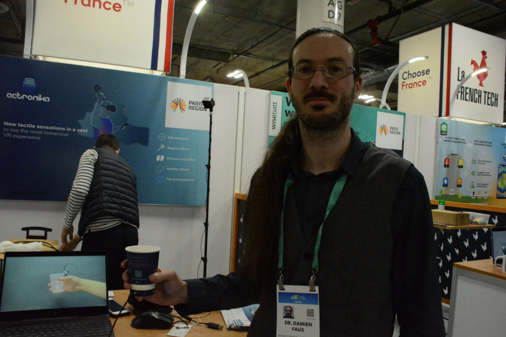 Dr Damien Faux holds a cup next to video showing how Actronika haptics platform can give it the "feel" of items dropping inside it at Eureka Park startup exhibit