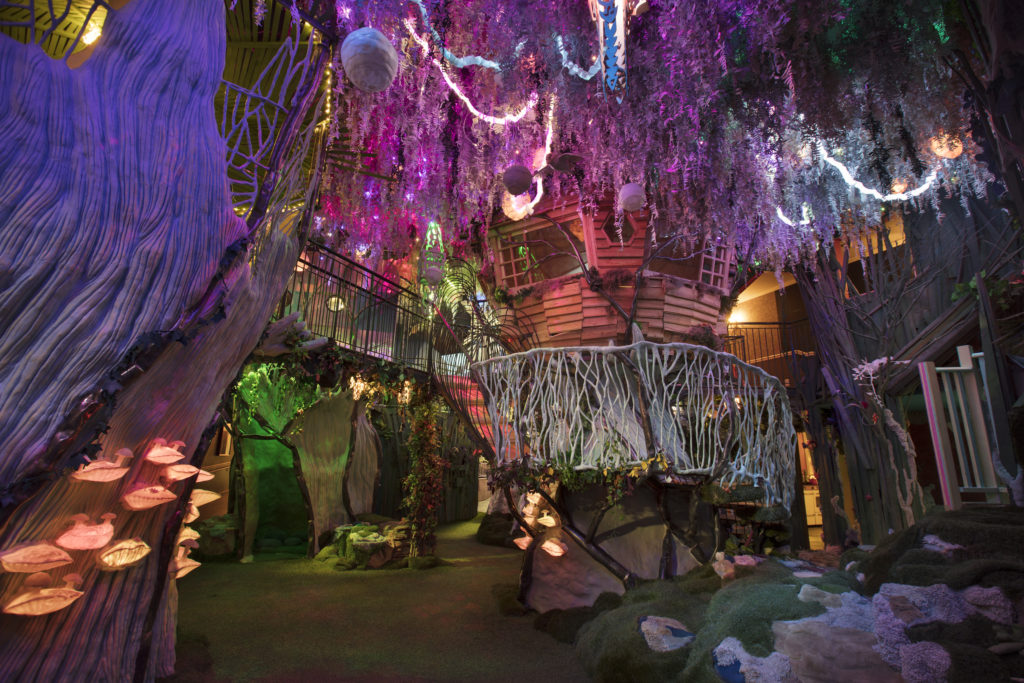 Wooden house perched atop a pedestal in the middle of a room, surrounded by multicolored lights, knotted ropes that look like Spanish moss, with circular orbs hanging from the ceiling over it, insie the Meow wolf installation