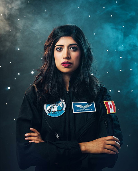 Dr. Shawna Pandya, in a blue flight suit with a Canadian flag decal and name badge, against a background of the Milky Way.
