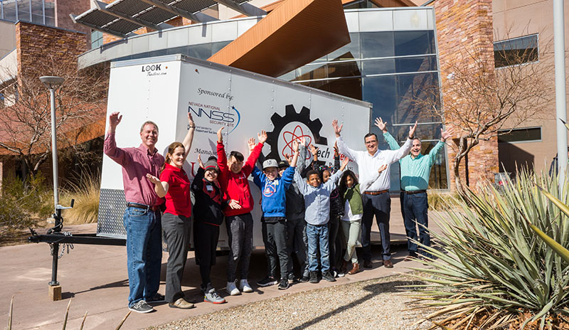 Tech Trekker staffers smile and line of elementary students smile and wave from in front of the Tech Trekker mobile lab outside the UNLV Engineering building
