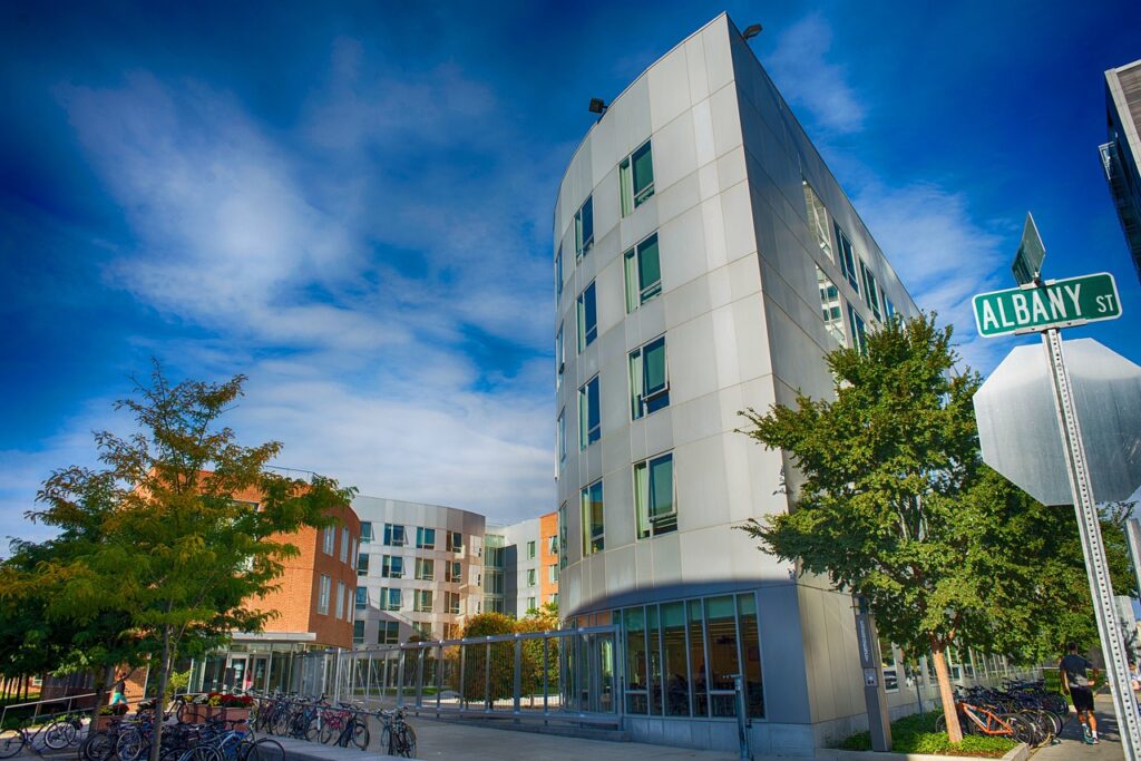 Campus at MIT with a tall gray four-story building and brick buildings in the distance.