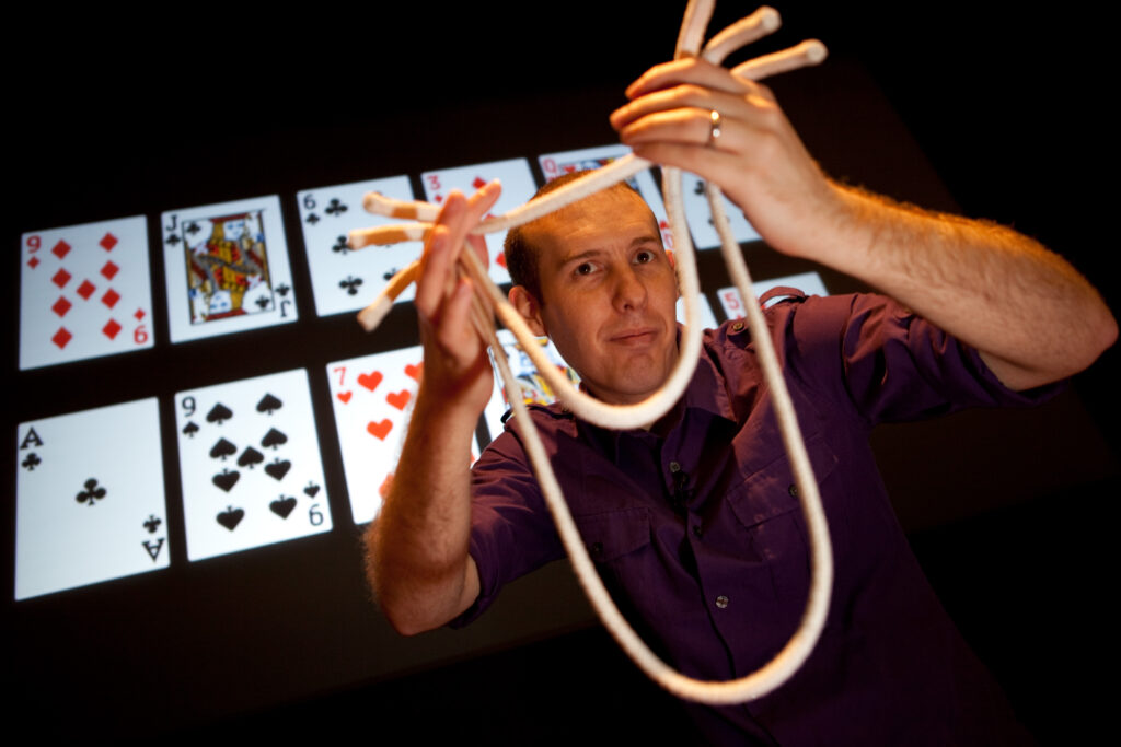 Science Magician Dr. Matt Pritchard looks through a knotted rope as he stands onstae with a background of nine playing cards projected on a screen.