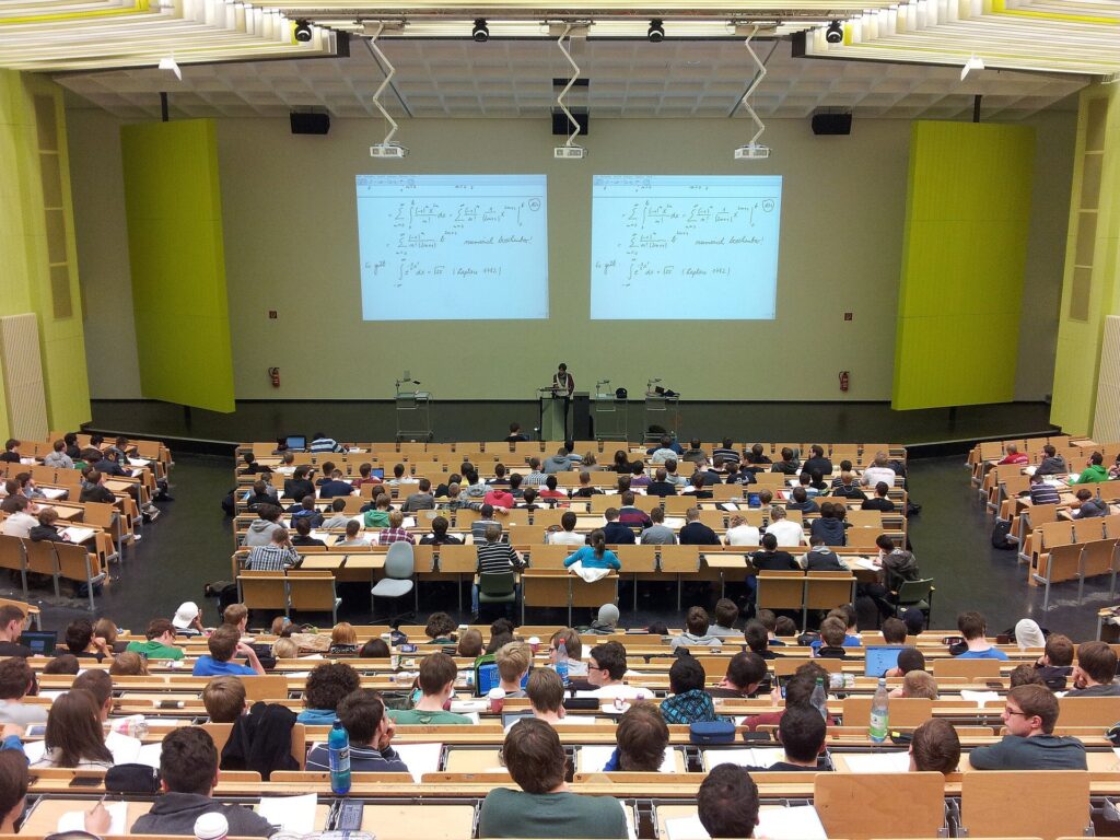 University students sit in a large lecture auditiorium with a lecturer standing before two screens at the front