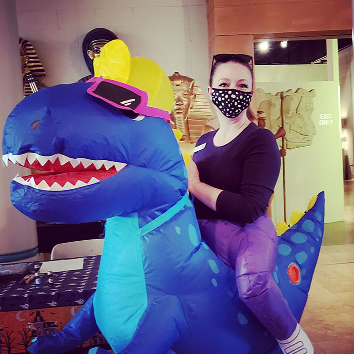 Kate Porter, in a black shirt and face mask, astride a blue "Dinosaur" ridable for "Dino-Ween".