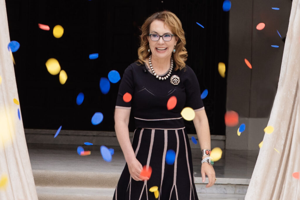 Medium shot of Gabby Giffords, wearing a black dress with a black-and-white striped skirt and silver jewelry including a large rose pin in a shower of multicolored rose petals