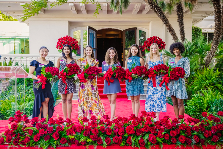 Seven members of Pasadena Tournament of Roses® 2023 Royal Court stand before Tournament House with bouquets of red roses in their arms.)