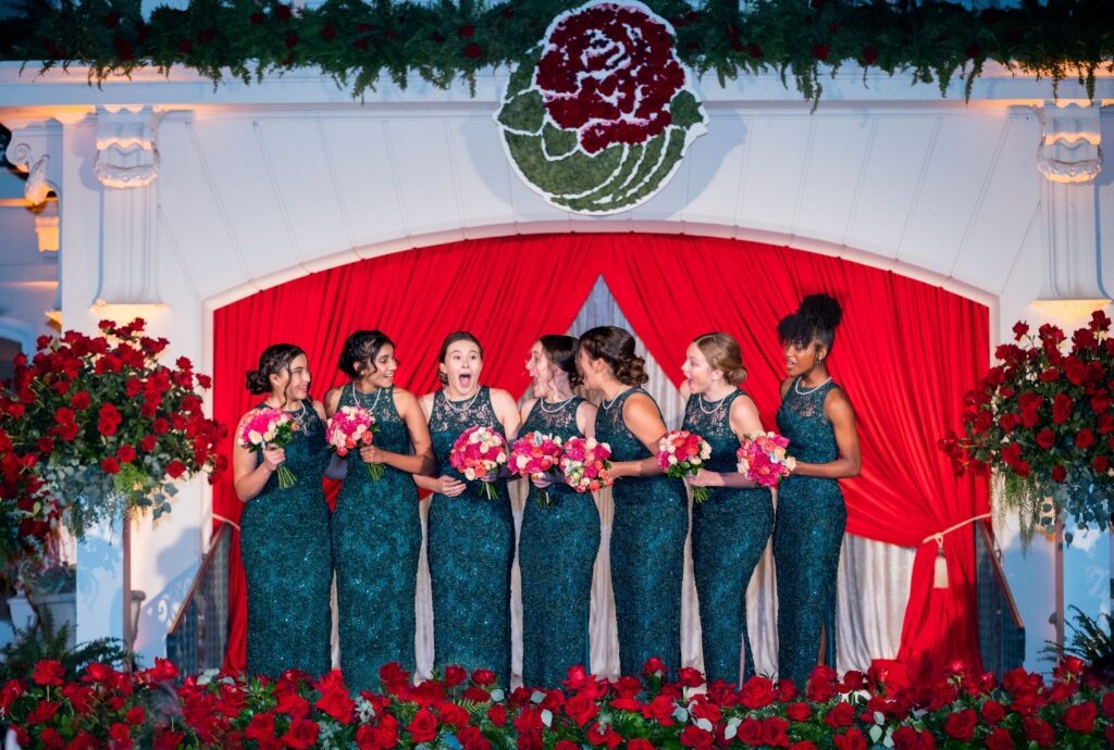 Members of the 2023 Royal Court, in sparkling black dresses, stand smiling with Bella Ballard, who's openmouthed with surprise after hearing herself announced as 2023 Rose Queen®, in front of a red curtain with a red rose medallion over it.
