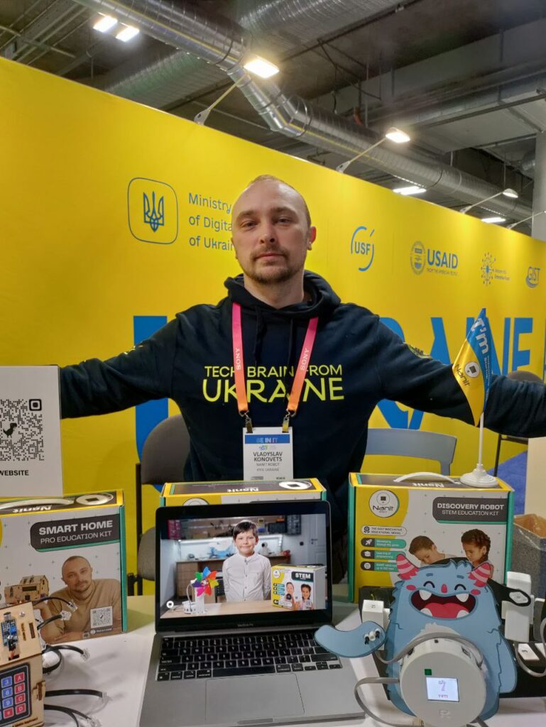 Nanit Robotics Founder and RoboHouse CEO Vladyslav Konov stands behind a display of robotic products including Smrt Home Pro Education Kit and Nanit Discovery Robot at CES 2023 display while wearing a black sweatshirt emblazoned with "Tech Brain from Ukraine", against a blue-and-yellow background.