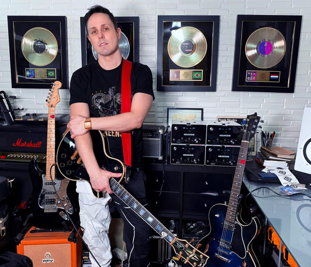 Meloscene Founder and CVO Chad Gerber, holding his guitar, stands in his studio with two guitars, a mixer and four gold and platinum records on the wall in the background.