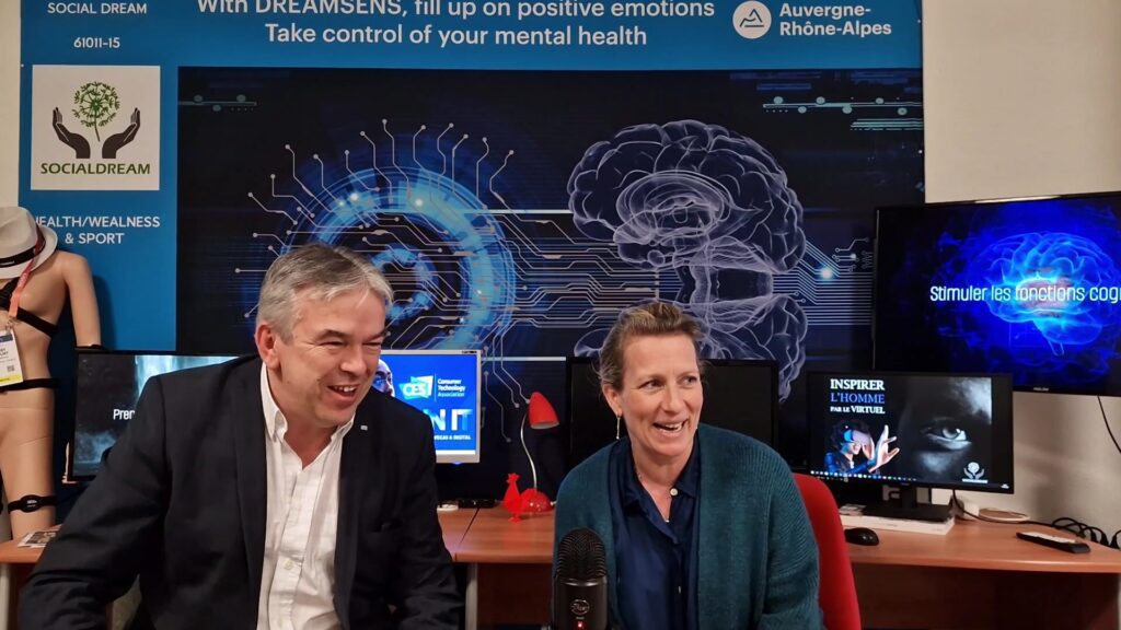 SAS SOCIALDREAM President Thierry Gricourt smiles as he sits with filmmaker/translator Ariane Tourneur in front of a display advertising his VR innovation with the words "Take control of your mental health".