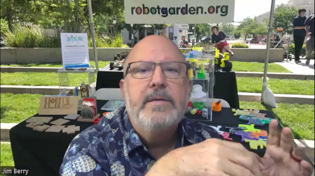 Robot Garden Treasurer and Head Maker Jim Berry discusses robots in front of a background showing his organization's outdoor booth, with banners that say "robotgarden.org" and "STEM".