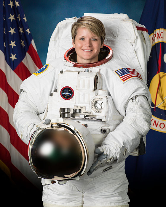 Astronaut Anne C. McClain smiles as she stands in her flight suit near the American flag.