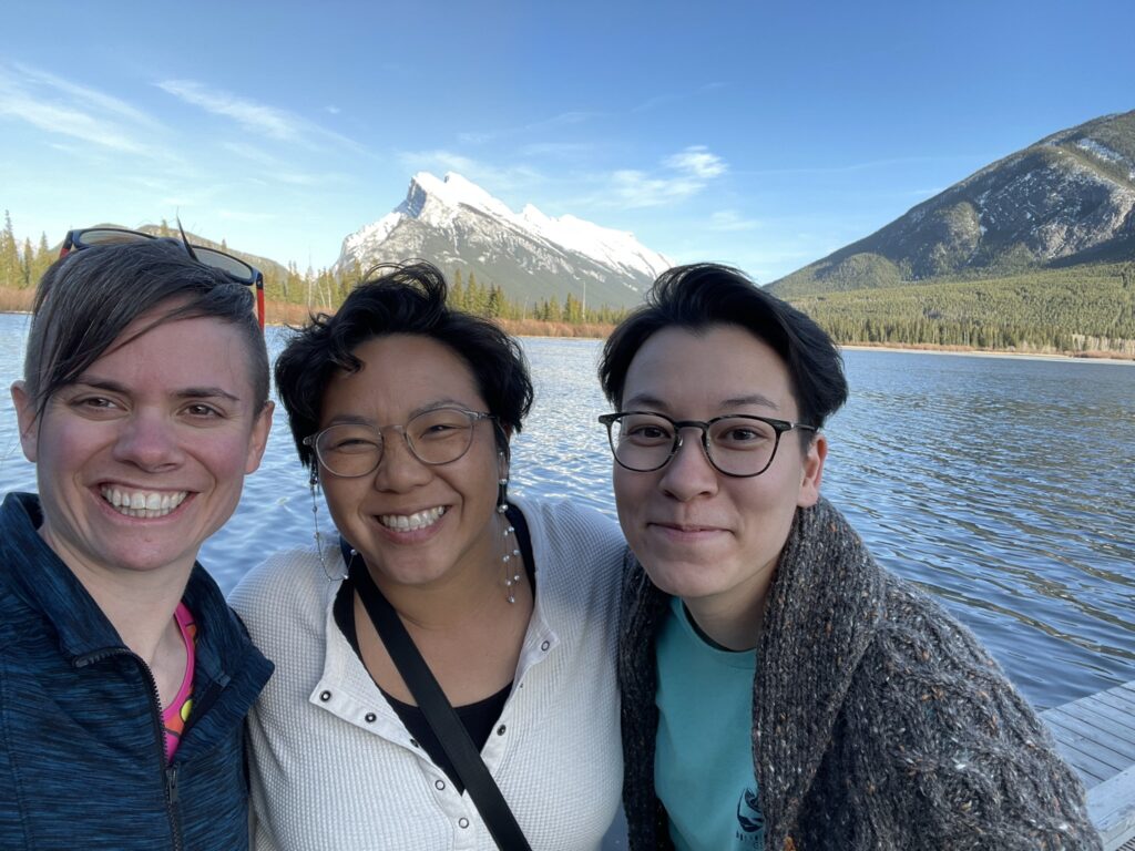 Found in the Fjords creators Dr. Louise Edwards, Tiffany Duong and Aya Walraven smile as they stand together before a Norwegian fjord against a bright blue sky.