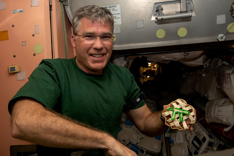 SpaceX Crew 6 Commander Stephen Bowen smiles aboard the ISS as he holds a lattice-crusted pie with a "PI" symbol on it. .