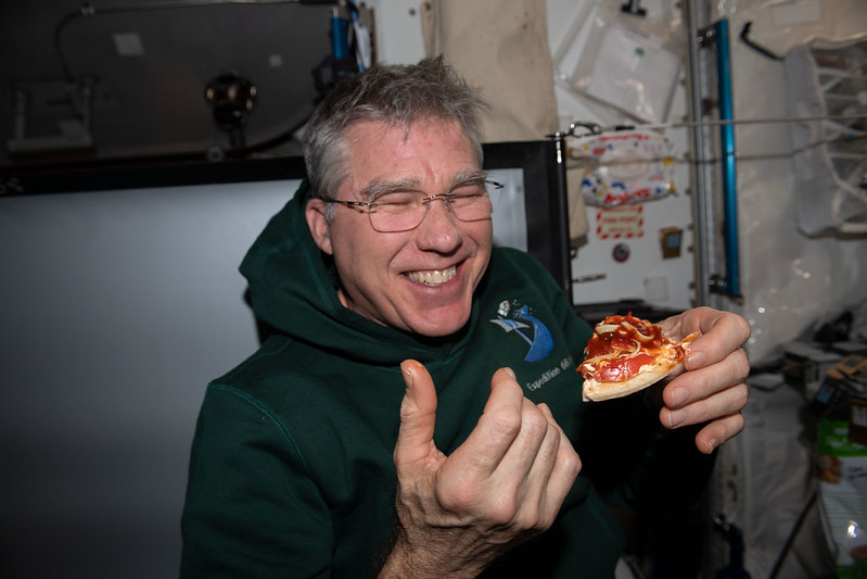 Steve, in a black hoodie, laughs as he enjoys a slice of pizza in zero gravity.