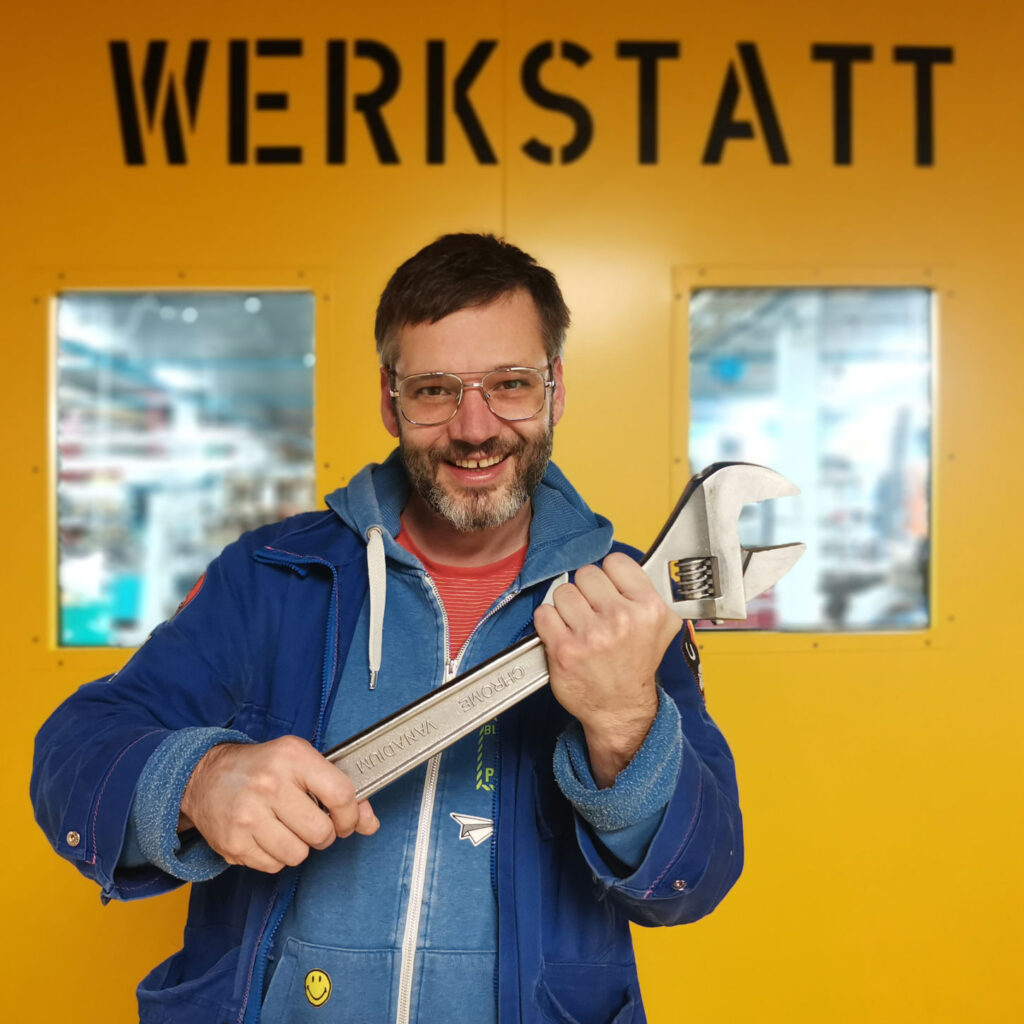 Artist and inventor wrench in front of a background with "WERKSTATT" printed on it in block letters.Niklas Roy smiles as he holds a wrench