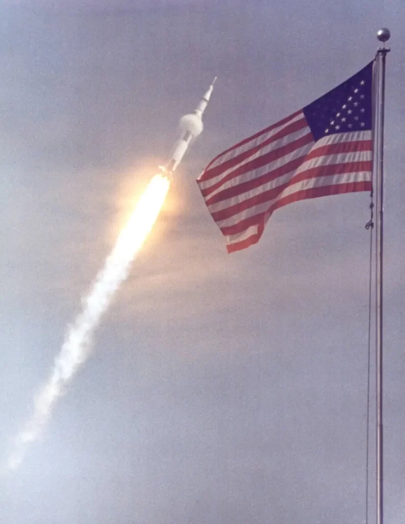 Apollo 11 blasts off for the Moon with an American flag in the foreground.