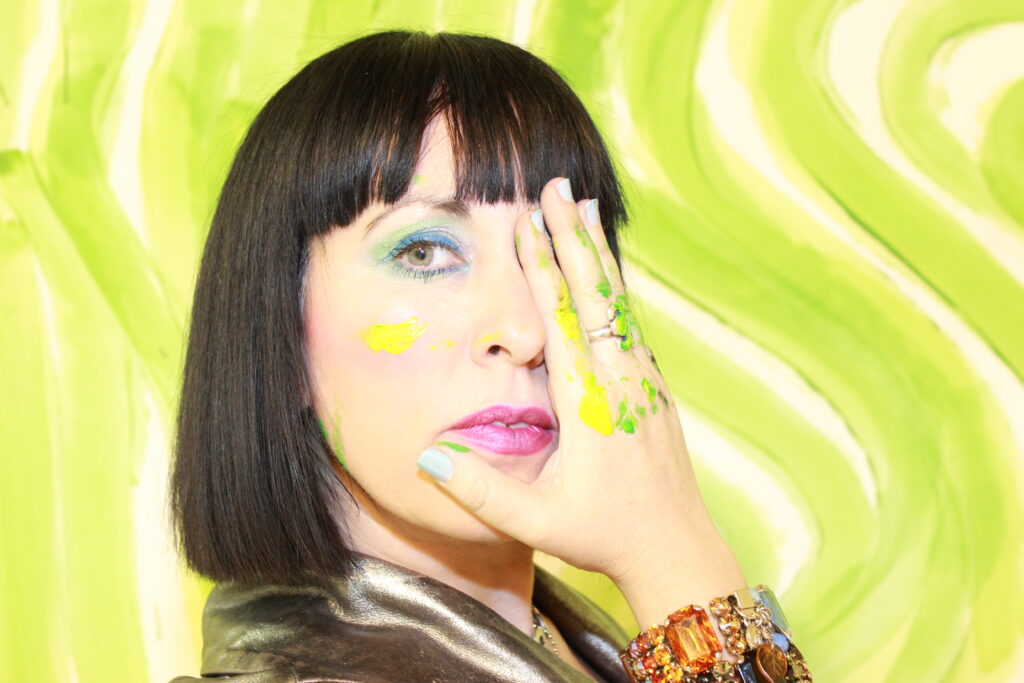 Fine artist and XR futurist Paige Dansinger, with paint splotches on her hand and cheek, playfully covers one eye as she poses in front of an abstract background.