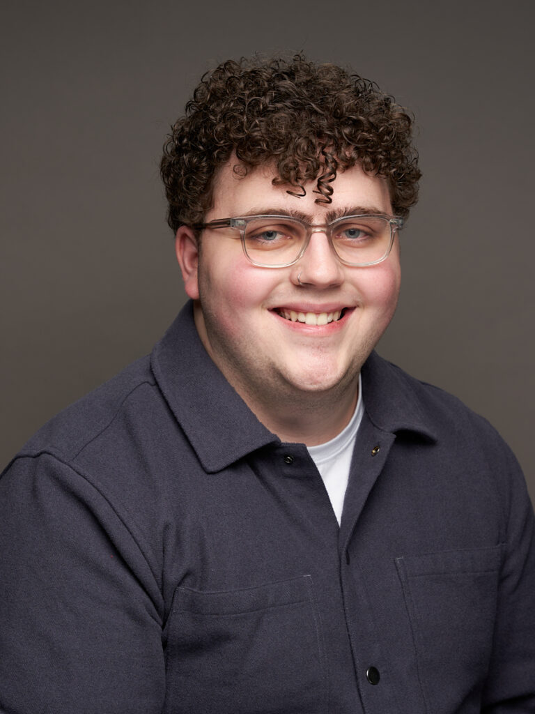 Accessible Gaming Lead, Neil Squire Society Makers Making Change Tyler Fentie smiles in a headshot. He's wearing a black open-neck shirt, has curly hair and glasses and is standing before a dark background.