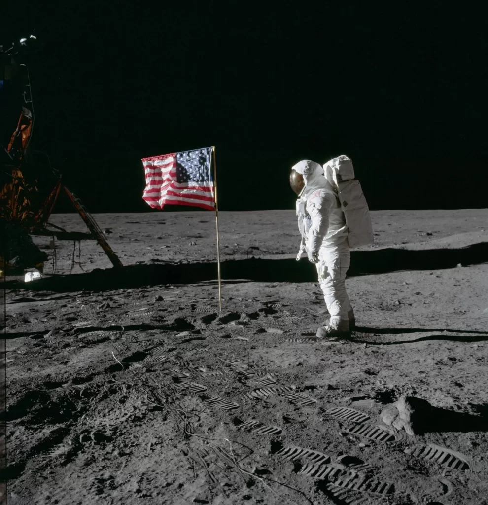 Neil Armstrong, in full space suit, walks on the lunar surface next to an American flag.