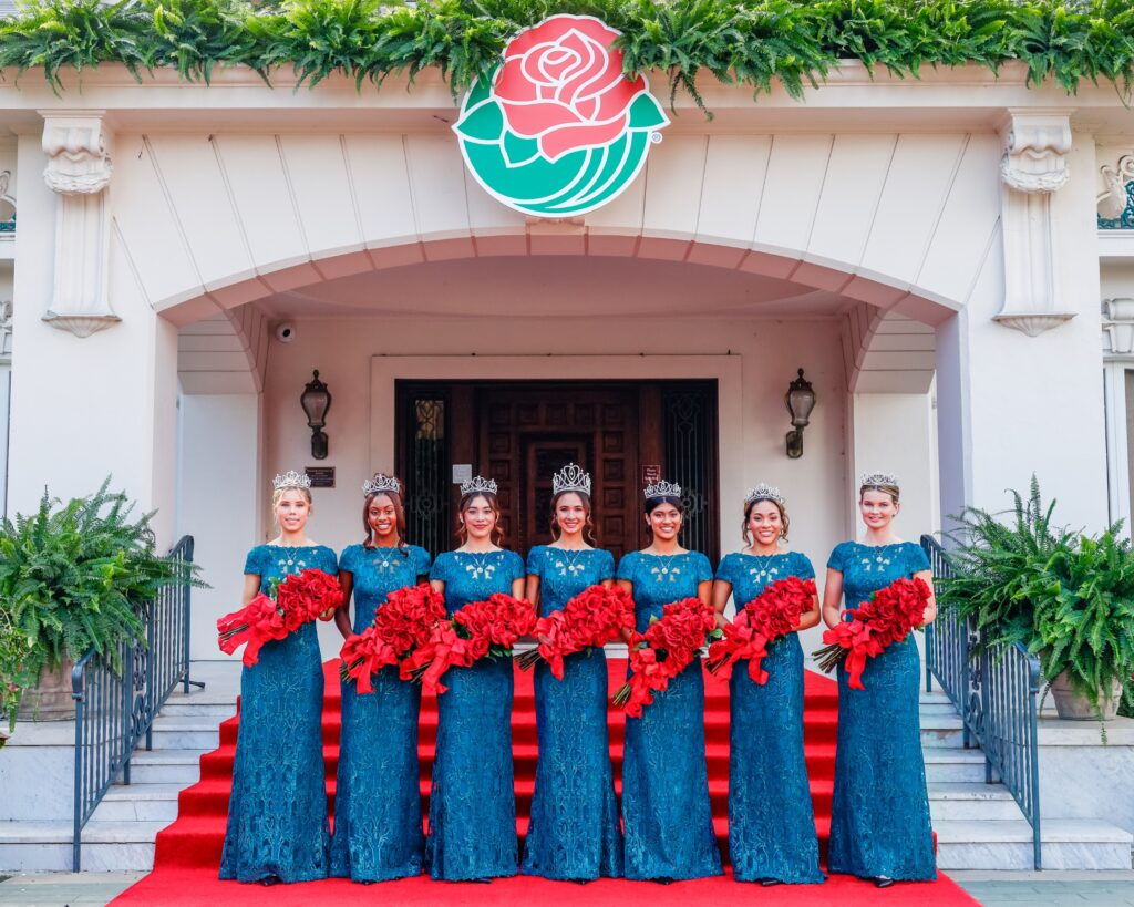 2025 Rose Queen Lindsay Charles, on the steps of Tournament House, flanked by her Rose Court, all wearing blue floor-length gowns and carrying red roses.