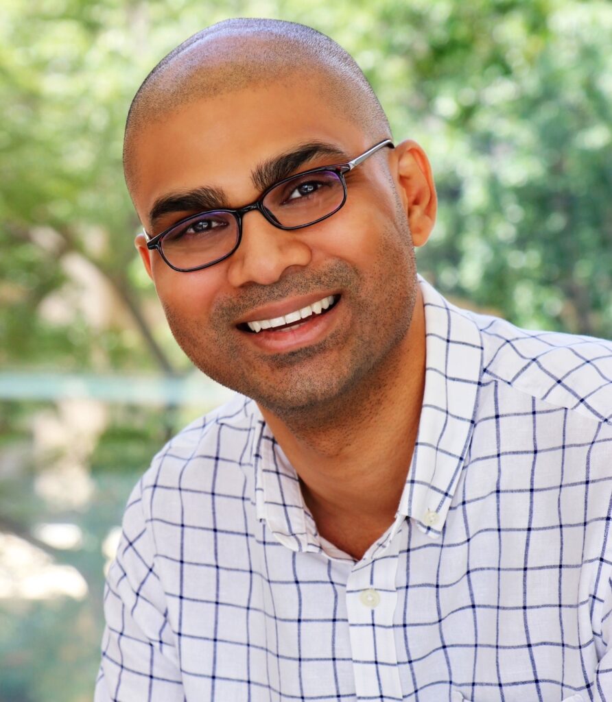 Dr. Cassini Nazir, in a checkered shirt, smiles against an outdoor background.