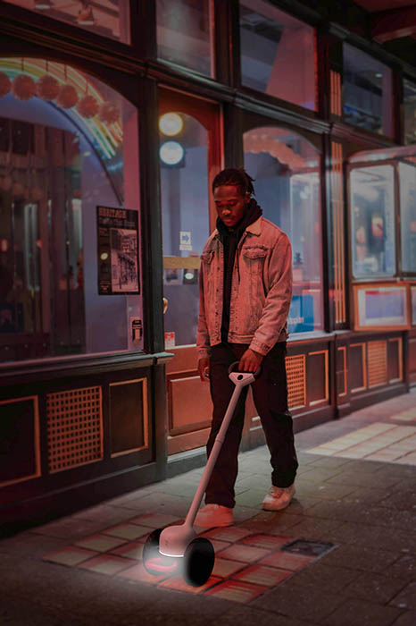 Smiling African-American man in a yellow jacket with black trim and black pants walks past lighted storefront at night, guided by Glide device in front of him.