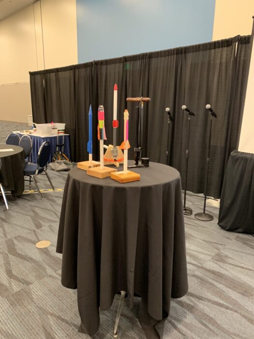 Three rockets from Air Rocket Works on a table in a conference room with three microphones and a curtain in the background, plus additional tables for attendees