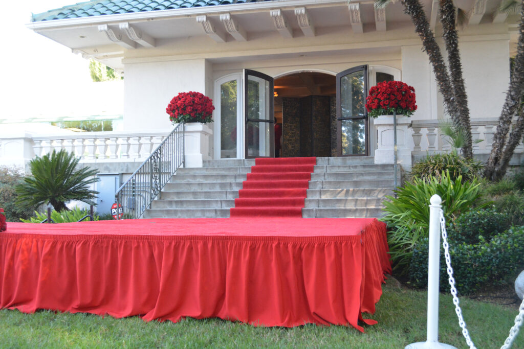 Open double doors of Tournament House with red roses flanking the opening and red carpet down the stairs to a red-carpeted platform.