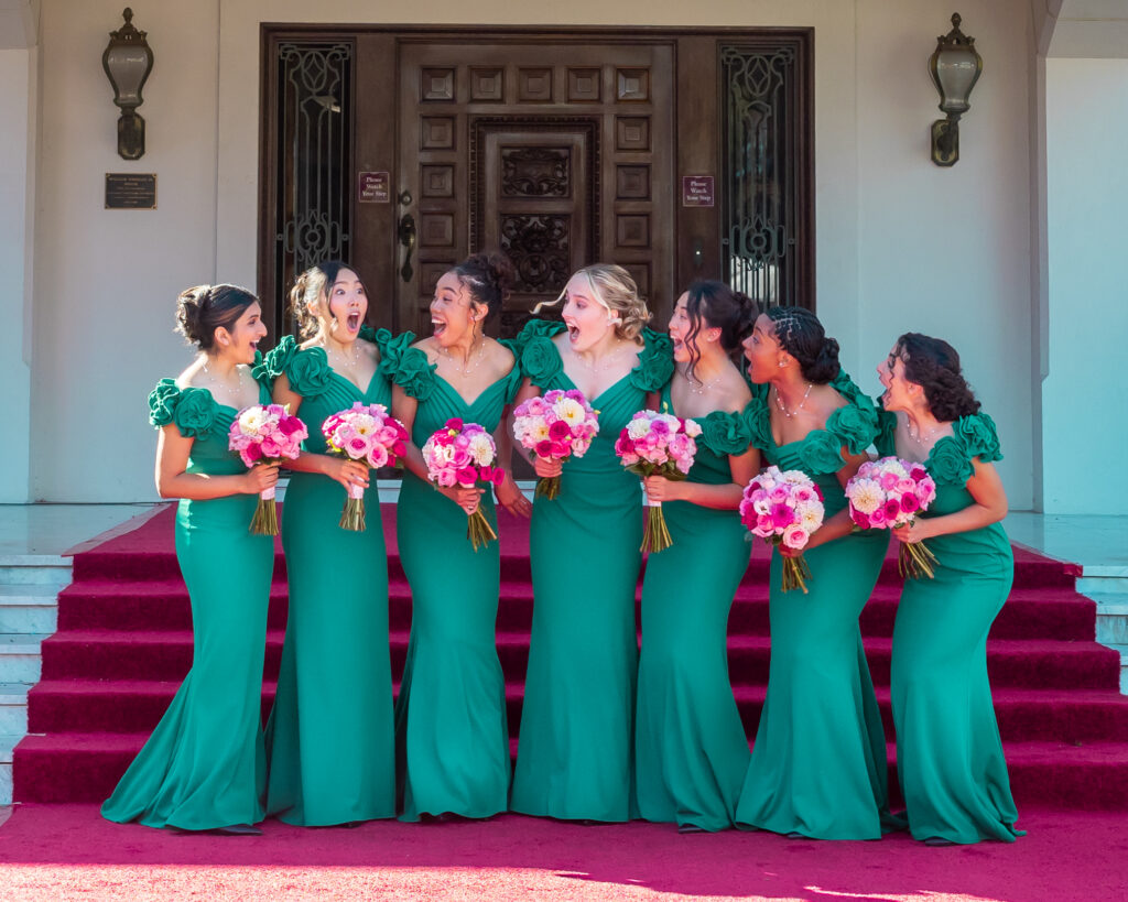 2026 Rose Queen Serena Guo stands openmouthed in surprise as six members of the Royal Court, smiling and wearing floor-length green gowns, face her with rose bouquets in their arms.