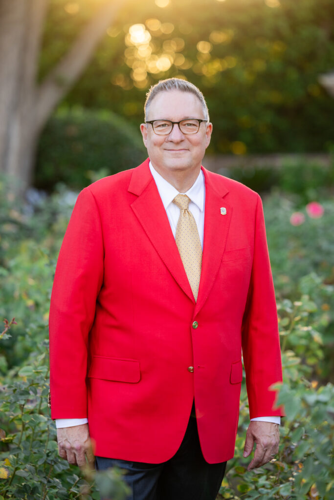 2027 Tournament of Roses President Terry Madigan, in a red jacket with a red striped tie, smiles against the background of the rose garden.