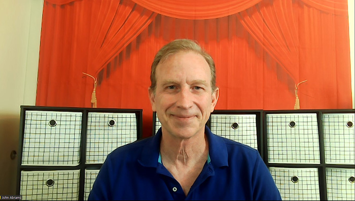 Amazing School Assemblies founder John Abrams smiles against a background of an orane theatrical curtain and black-and-white screens.