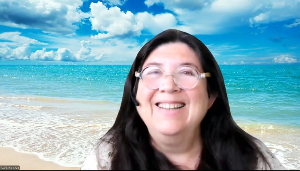 VR pioneer Dr. Carolina Cruz-Neira smiles against a Zoom background of a Florida beach with blue sky and puffy white clouds