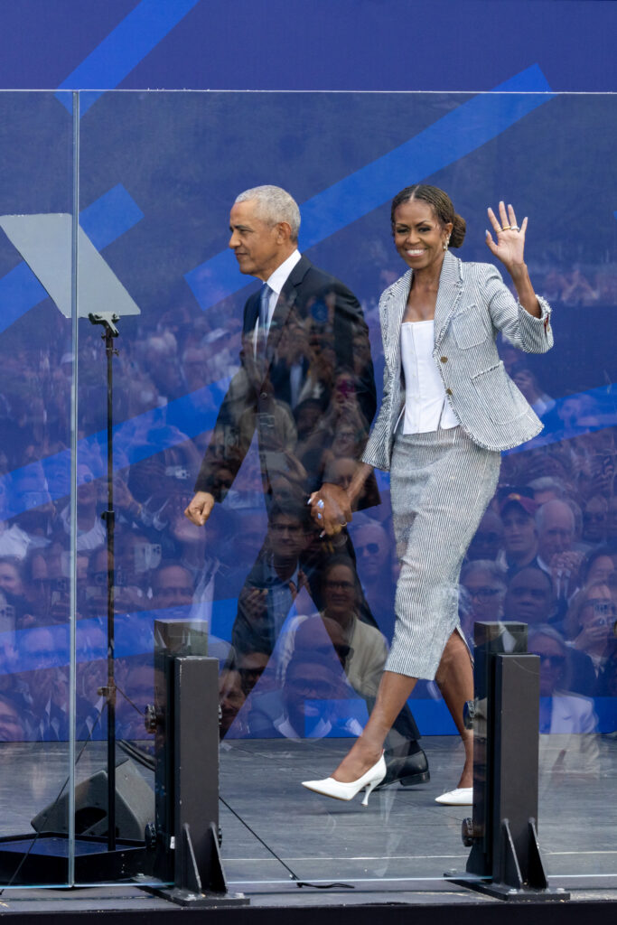 President Barack Obama and Mrs. Michelle Obama, holding hands, walk onstage as she waves to the audience.